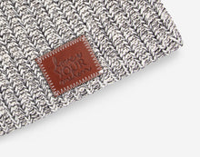 Load image into Gallery viewer, This black speckled beanie features a red maple leather patch debossed with the Love Your Melon logo on the front. These beanies are very cozy and warm and have a slouchier fit. Details: Yarn Color: Natural & Black Sizing: 9” x 11” standard when uncuffed. One Size. Content: 99% Cotton 1% LYCRA® Care: Machine wash cold. Use mild detergent. Tumble dry on high.