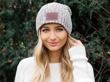 Load image into Gallery viewer, This black speckled beanie features a red maple leather patch debossed with the Love Your Melon logo on the front. These beanies are very cozy and warm and have a slouchier fit. Details: Yarn Color: Natural & Black Sizing: 9” x 11” standard when uncuffed. One Size. Content: 99% Cotton 1% LYCRA® Care: Machine wash cold. Use mild detergent. Tumble dry on high.